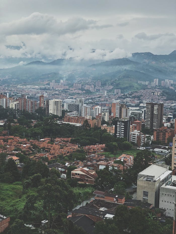 Aerial view of a cityscape with urban buildings and mountains in the background, under a cloudy sky.
